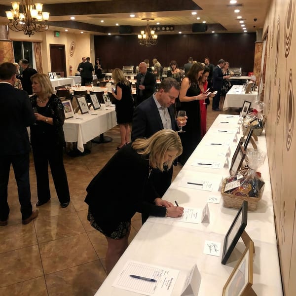 Guests in formal attire mingle and sign bid sheets at a silent auction event in a large room.