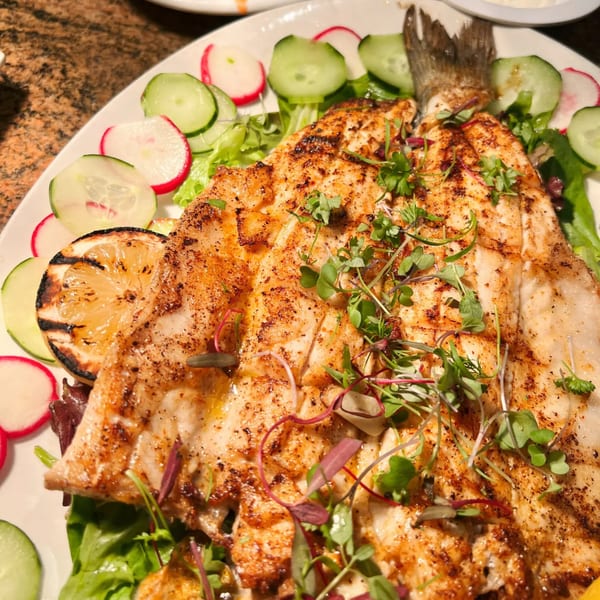 Whole grilled fish, garnished with microgreens, grilled lemon, sliced radishes, and cucumbers on a bed of lettuce.